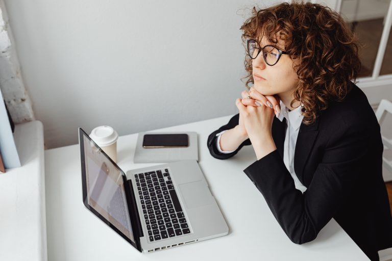 A pensive woman in business attire sits at a desk with a laptop, contemplating work