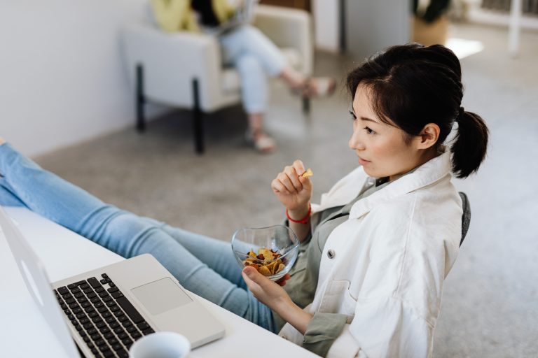 Young woman enjoying a snack while working on a laptop in a casual office setting.