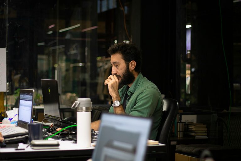 A man sits in front of his desk at his office late at night and has an exhausted expression on his face